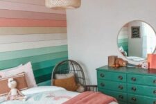 a bright rainbow teen girl room with a bold rainbow paneled wall, a bed with bright bedding, an emerald dresser, a round mirror and a woven pendant lamp
