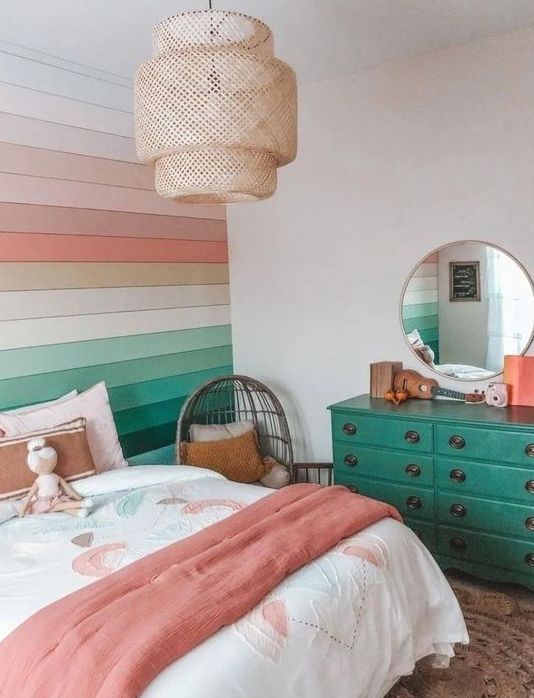 a bright rainbow teen girl room with a bold rainbow paneled wall, a bed with bright bedding, an emerald dresser, a round mirror and a woven pendant lamp