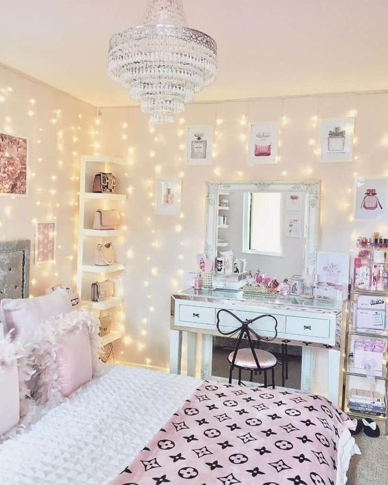 a glam teen bedroom with a grey bed with pink and blue bedding, a white desk and a mirror, a crystal chandelier and a shelf in the corner