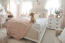 a neutral and lovely teen girl’s bedroom with plaid wallpaper, a white bed with neutral bedding, white dressers, a blush vanity and a large mirror, a sofa