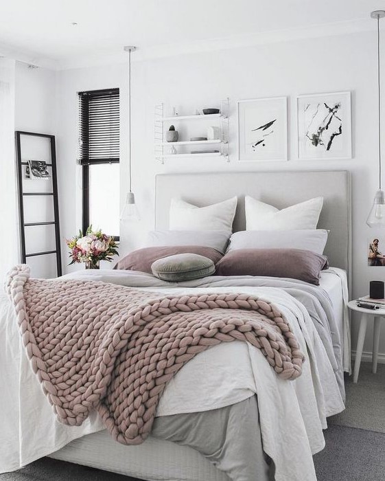 a neutral and pastel bedroom with a grey upholstered bed, layered blankets, black touches and bulbs hanging down