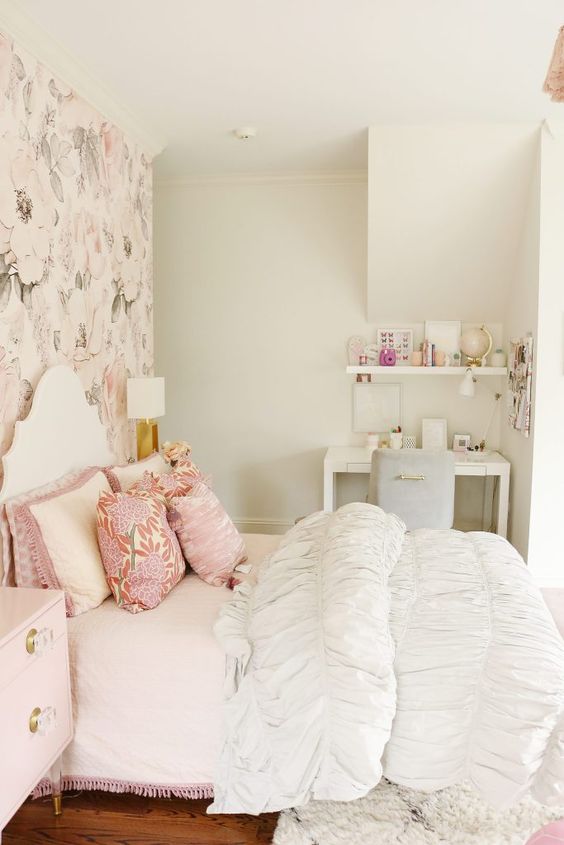 a teen girl bedroom in neutrals and pastels, with a floral accent wall, a white bed with printed and neutral bedding, a blush nightstand, a white desk and a grey chair