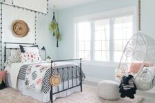 an airy boho teen girl bedroom with light blue walls, a bed with a cnaopy, printed bedding, a suspended chair and a blush rug