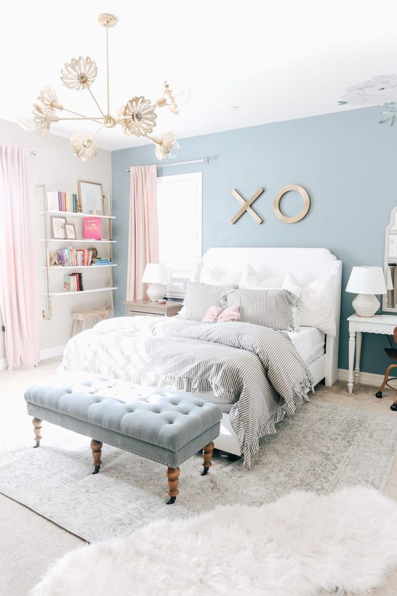 an airy teen girl bedroom with an ocean blue accent wall, open shelves in the corner, a white bed with neutral bedding, a powder blue upholstered bench