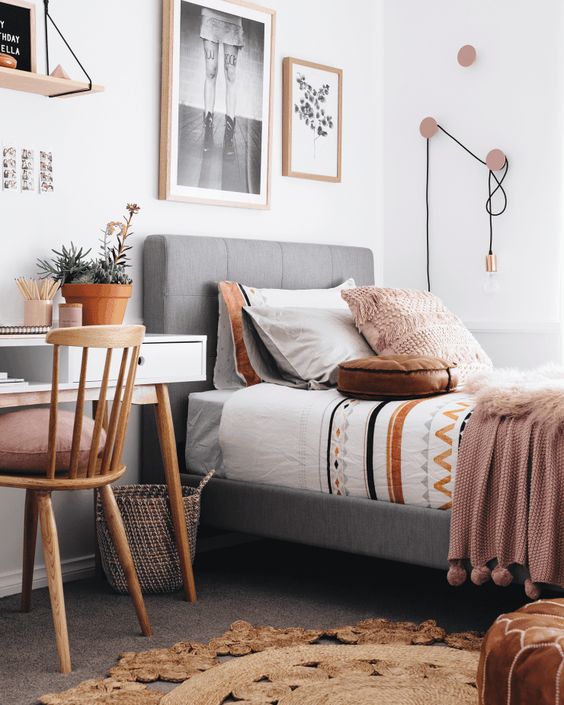 a boho Scandi bedroom with a grey upholstered bed, printed and pastel bedding, a desk and a stained chair, a mini gallery wall