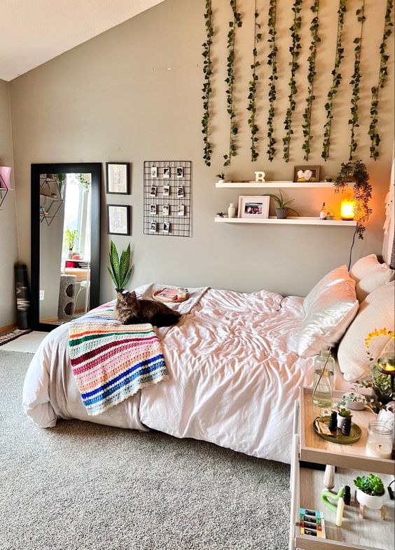 a neutral boho bedroom with greige walls, a bed with neutral bedding, open shelves, hanging greenery, a nightstand with blooms