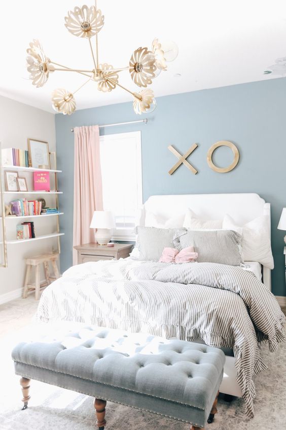 a pastel teen bedroom with a blue accent wall, a white bed with printed bedding, a blue upholstered bench, open shelves, a gold chandelier