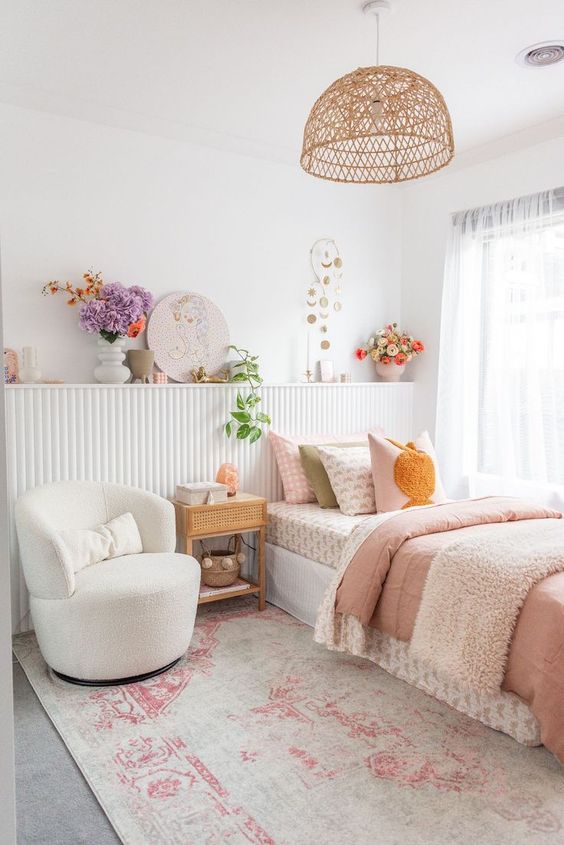 a romantic girl's bedroom with a paneled shelf with decor and blooms, a bed with pastel bedding, a creamy chair and a woven pendant lamp