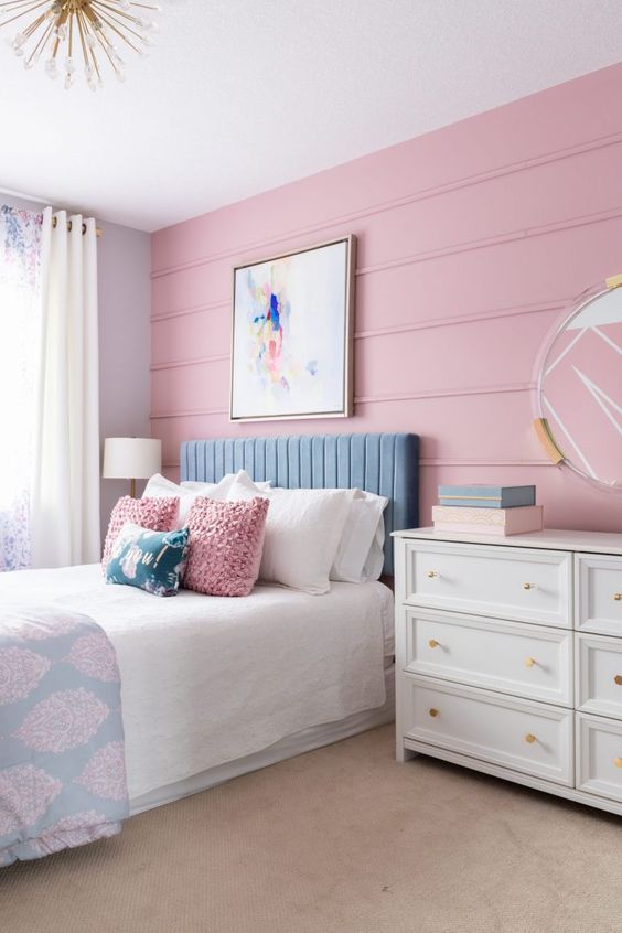 a stylish pastel teen bedroom with a pink panel accent wall, a blue upholstered bed, a white dresser, a mirror and a sunburst chandelier