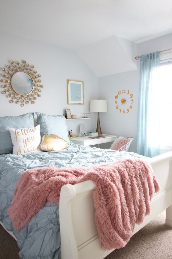 a welcoming teen bedroom with a small vanity and desk, a white bed with blue bedding, blue curtains, a sunburst mirror