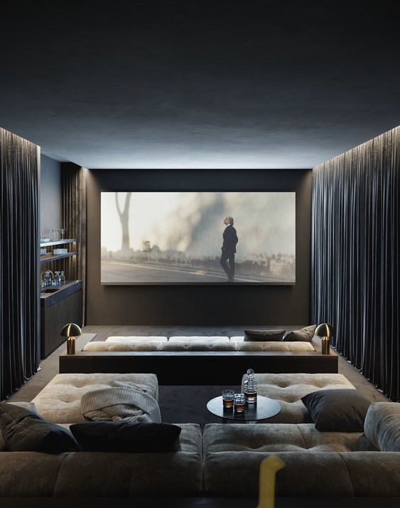 a chic moody home theater with curtains, a large screen, grey seating furniture, neutral ottomans and a small home bar in the corner