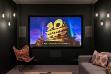a laconic and stylish dark home theater with black walls, a grey corner sofa, a wooden coffee table, a butterfly chair and a large screen