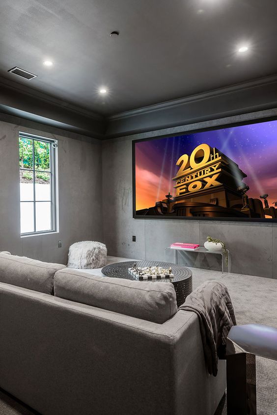 a modern home theater with concrete walls, grey seating furniture, a large screen, an acrylic console table and grey textiles