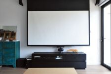 a simple and laconic home theater with a glazed wall, a screen, a black media console, a stained coffee table and a grey sofa plus lots of lights