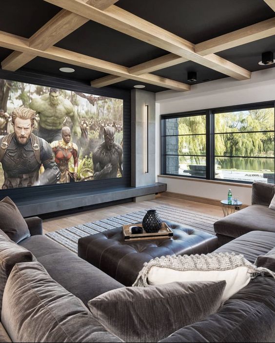 a welcoming home theater with a large window, a grey sofa, a leather ottoman, a large screen and a paneled ceiling is cool