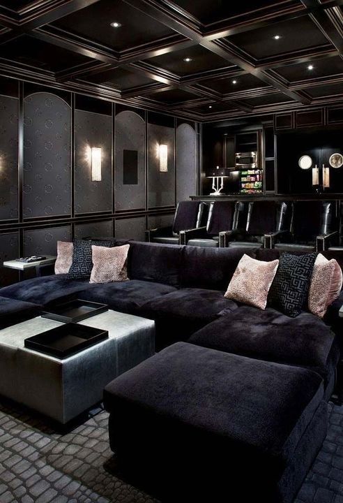 an elegant moody home theater in black, with a large sofa and lots of comfrotable chairs, a pouf and pink pillows, built-in lights