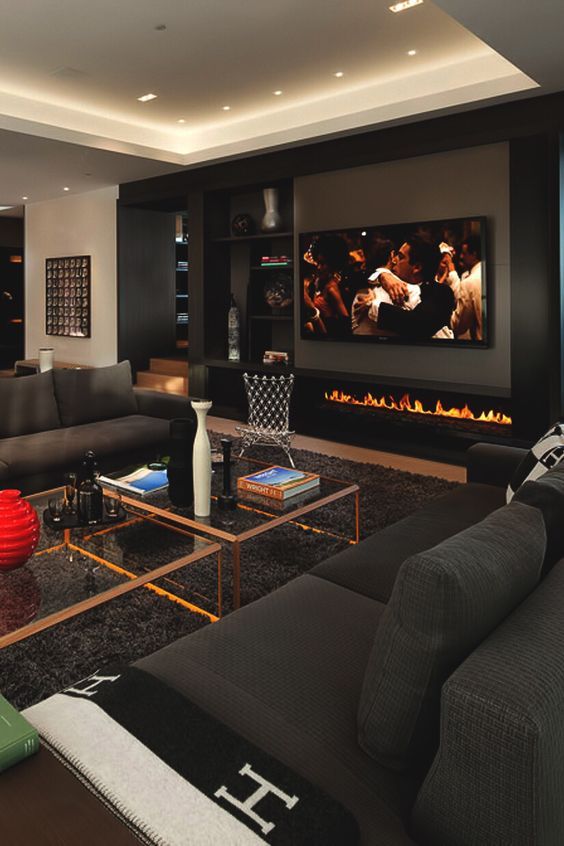 an elegant moody living room that doubles as a home theater, with a large screen and a built-in fireplace, black sofas, glass coffee tables and built-in lights