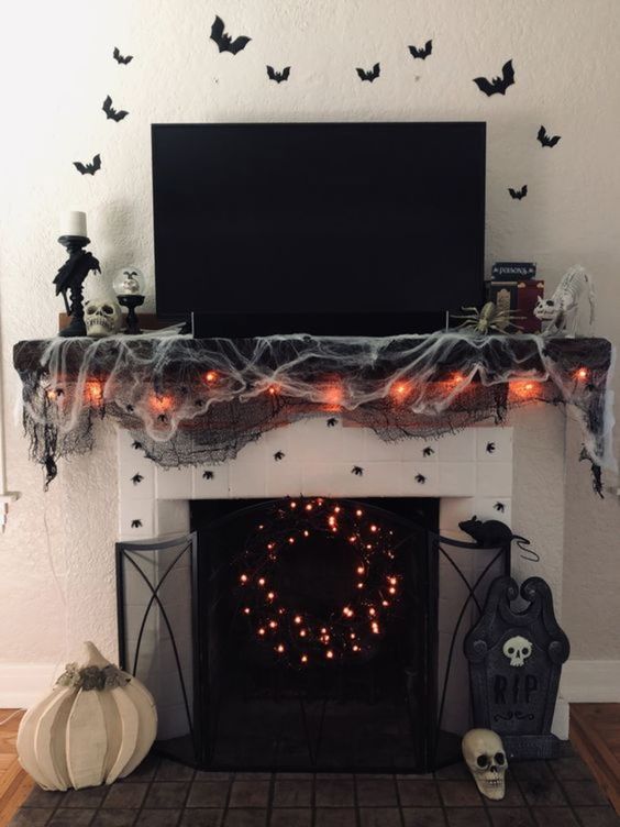 a black Halloween mantel with black and white spider web, lights, spiders, a lit up wreath and candleholders plus bats over the mantel