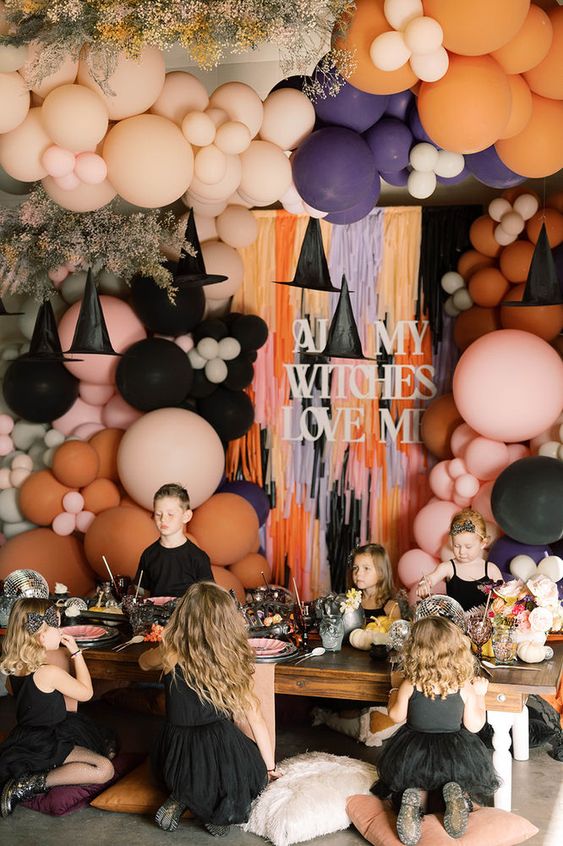 a gorgeous colorful Halloween party space with colorful balloons, fringe, witches' hats, a low table with disco balls and pink and black plates