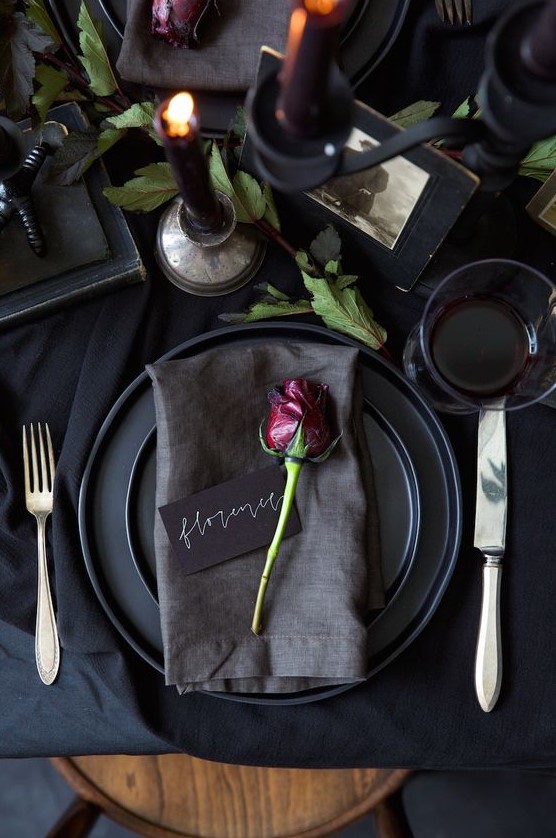 a refined black Halloween table setting with black linens, plates, candles, vintage books and photos, greenery and simple cutlery