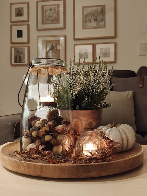 a Thanksgiving arrangement of a wooden tray, a faux pumpkin, dried leaves, a candle lantern, another one filled with acorns and some blooms