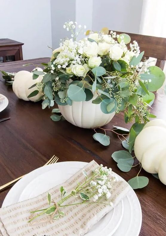 a beautiful and delicate fall centerpiece of a white pumpkin, white blooms and greenery plus some white pumpkins on the table