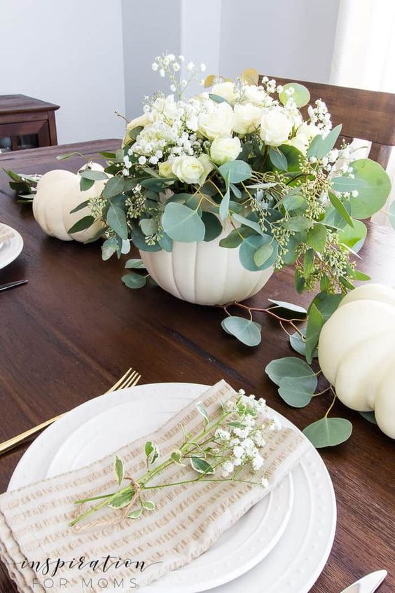 a beautiful and delicate fall centerpiece of a white pumpkin, white blooms and greenery plus some white pumpkins on the table