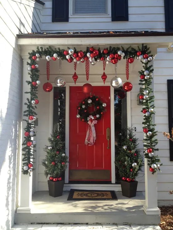 a bold Christmas porch with red and silver ornaments, fir garlands, a wreath with ornaments and mini Christmas trees in pots