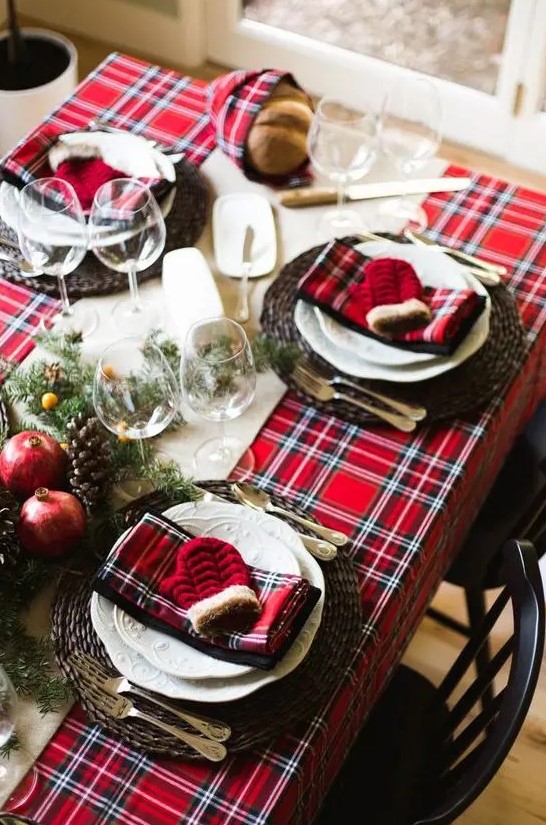 a bold Christmas table setting with a red plaid tablecloth and napkins, woven chargers, white porcelain, fir branches, berries, pomegranates and pinecones