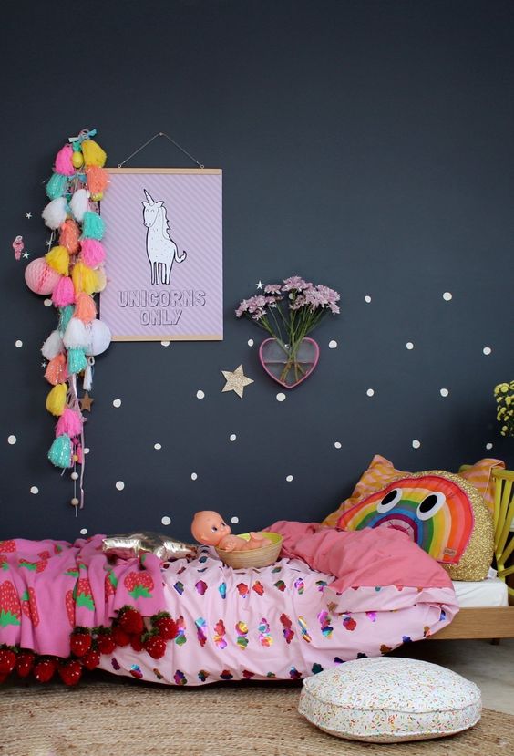 a bold and dreamy kid's room with a black accent wall, a bed with colorful bedding, some tassels and pompoms, blooms and art