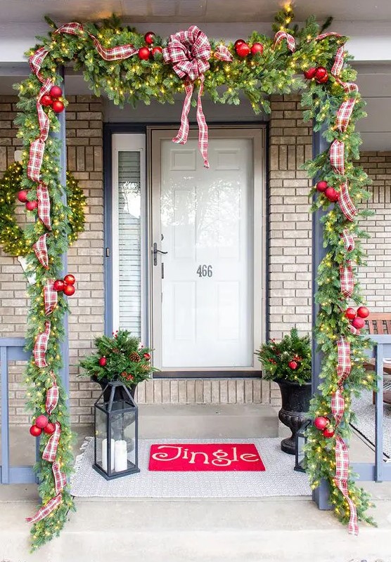 a bright Christmas porch with an evergreen garland with lights, red ornaments, plaid ribbons and bows, a red rug, candle lanterns and arrangements