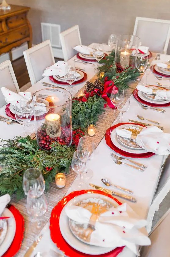 a bright and chic Christmas table setting with evergreens, red berries, candles wrapped with bark, red chargers and polka dot napkins