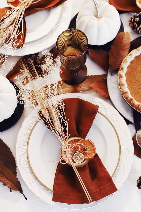 a bright boho Thanksgiving place setting with white plates, a rust-colored napkin with dried citrus slices and wheat