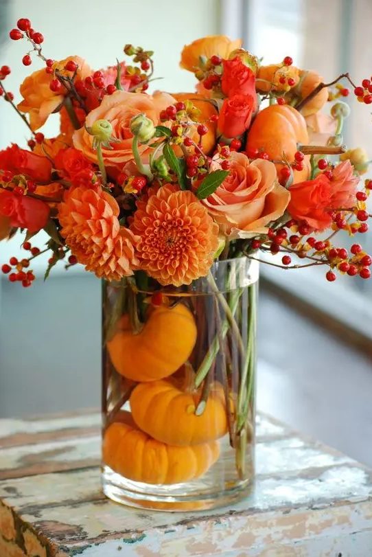 a bright fall or Thanksgiving centerpiece of a clear vase with orange gourds, bold orange blooms and berries is a very fresh and vibrant idea