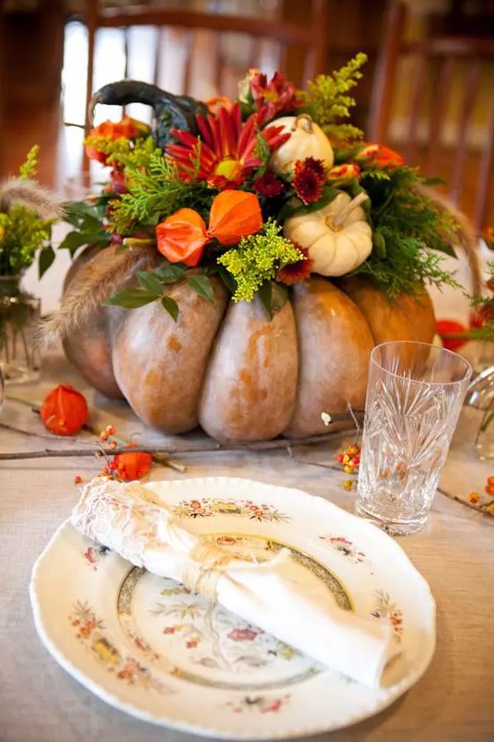 a bright rustic Thanksgiving centerpiece of a heirloom pumpkin with gourds, dired and bold blooms, greenery, wheat and blooming branches