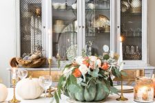 a chic Thanksgiving centerpiece of a green pumpkin, blush and orange blooms and lots of greenery, gold candleholders and tall and thin candles