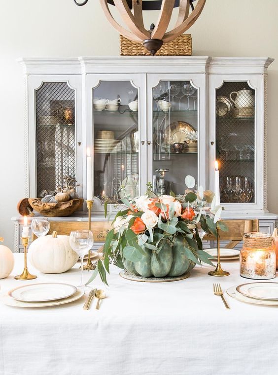 a chic Thanksgiving centerpiece of a green pumpkin, blush and orange blooms and lots of greenery, gold candleholders and tall and thin candles