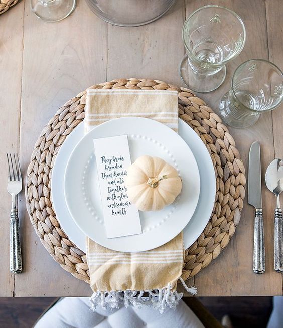 a chic neutral Thanksgiving place setting with a wovne placemat, white porcealin, a tan npkin and pumpkin plus a card