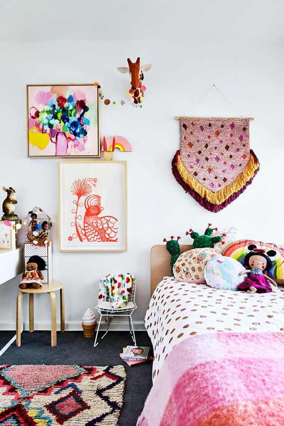 a colorful and fun boho-inspired kid's room with colorful bedding and pillows, a colorful gallery wall with a hanging and some stools and rugs