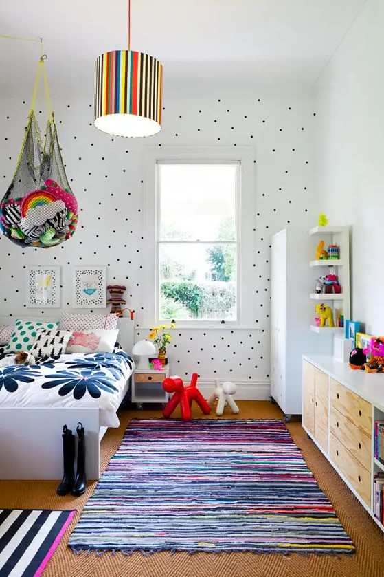 a colorful kid's room with a polka dot wall, colorful bedding and rugs, bold toys in nets over the bed