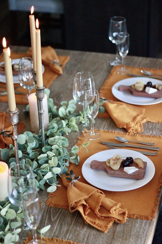 a delicate Thanksgiving place setting with a rust-colored placemat and a matching napkin, a white plate and a wooden board with some appetizers