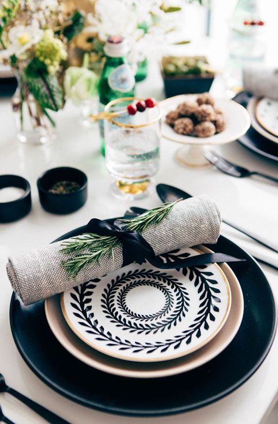 a large black plate, a gilded one, a black and white printed plate on top and a burlap napkin with greenery for a chic Thanksgiving table