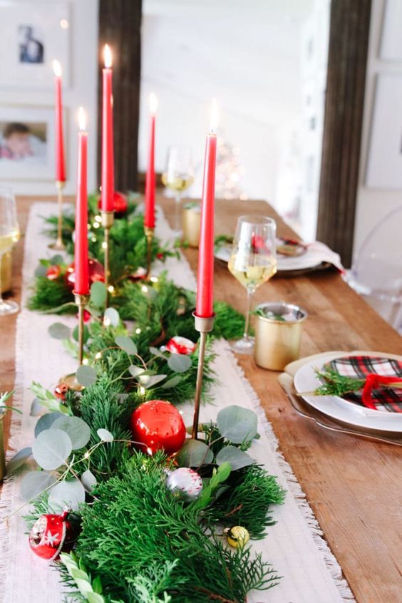 a lovely Christmas tablescape with an evergreen runner, silver, gold and red ornaments, red thin and tall candles and plaid plates