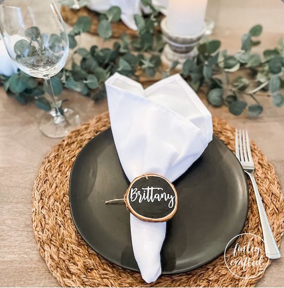 a lovely rustic Thanksgiving place setting with a woven placemat, a black plate, a white napkin, a chalk board wood slice as a place card