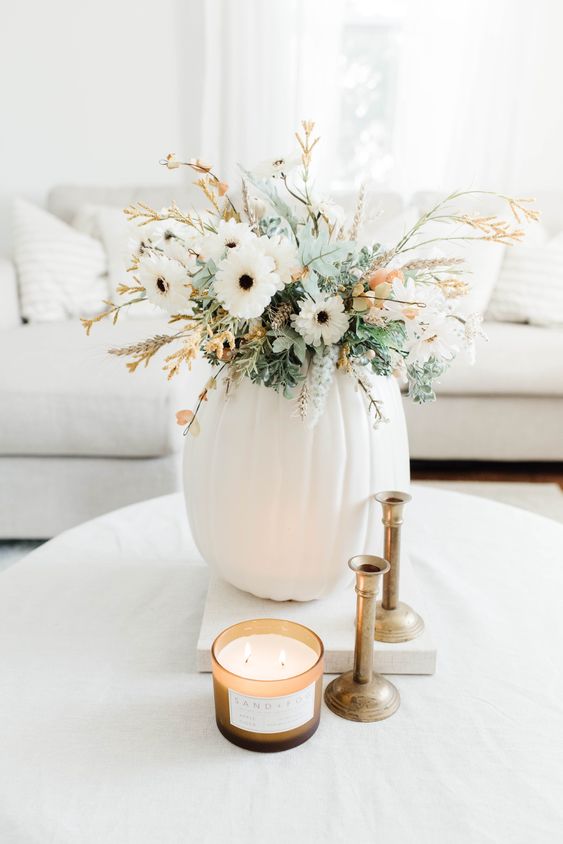 a neutral Thanksgiving centerpiece of a white pumpkin, white blooms, greenery and some blooming branches is cool