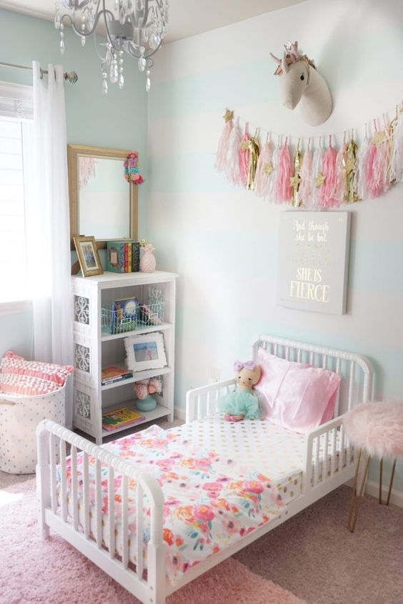 a pastel kid's room with light green walls, a white bed with colorful bedding, a white storage unit and colorful tassels