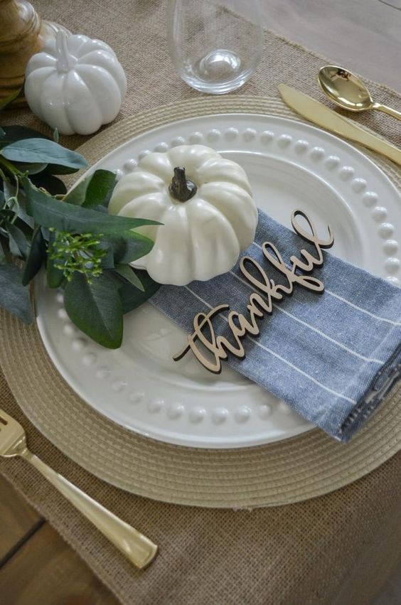 a pretty and natural Thanksgiving place setting with a woven charger, a plate, a faux pumpkin, a blue napkin and greenery