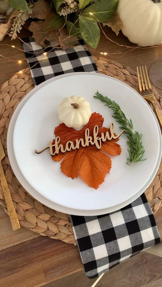 a pretty modern farmhouse tablescape with a woven placemat, white porcelain, a fall leaf, a pumpkin, greenery and calligraphy plus lights