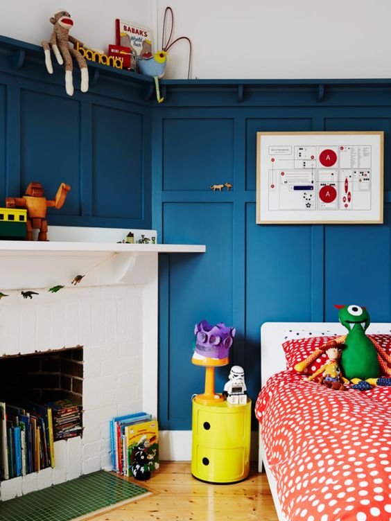 a quirky kid's room with blue paneling on the walls, a white birkc fireplace used for book storage, a bed with colorful bedding and toys