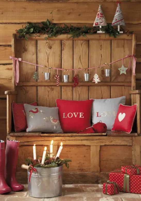 a rustic wooden bench decorated with grey and red printed Christmas pillows, with a grey and red garland, red gift boxes and a bucket with candles